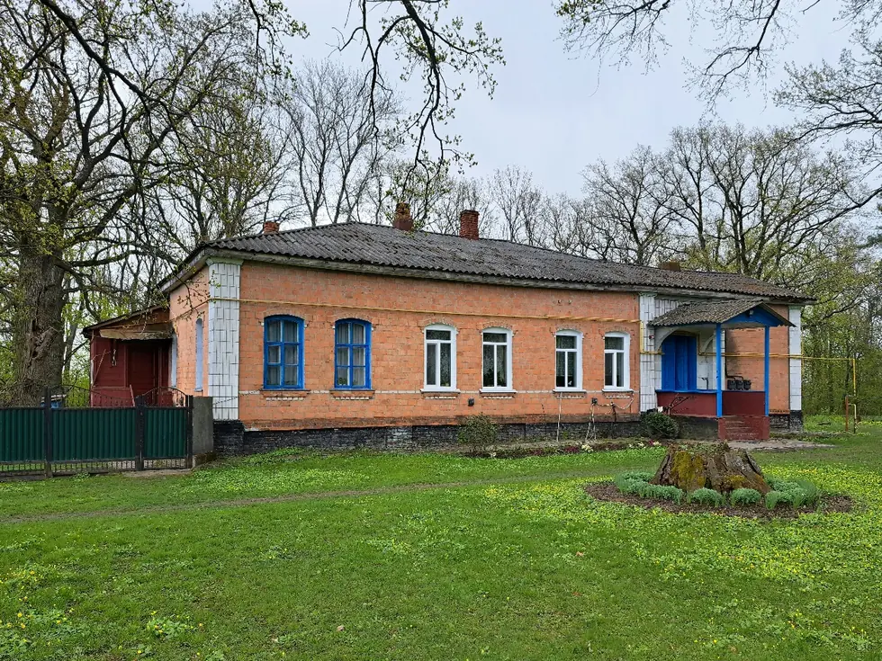 Довгий одноповерховий будинок із синім ґанком серед дубів на зеленій галявині
