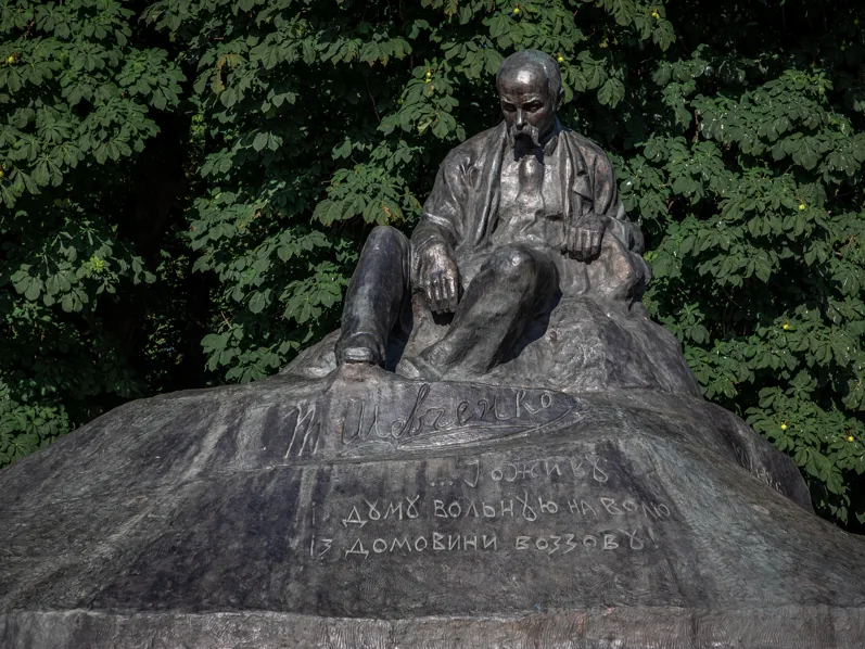 Бронзова скульптура Тараса Шевченка, який сидить на камені, серед зелених дерев